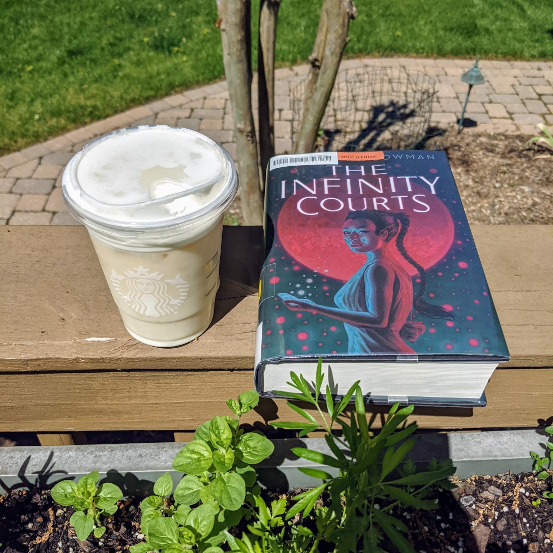 Iced green tea Starbucks drink sits next to a library hardcover copy of The Infinity Courts, in front of a herb garden