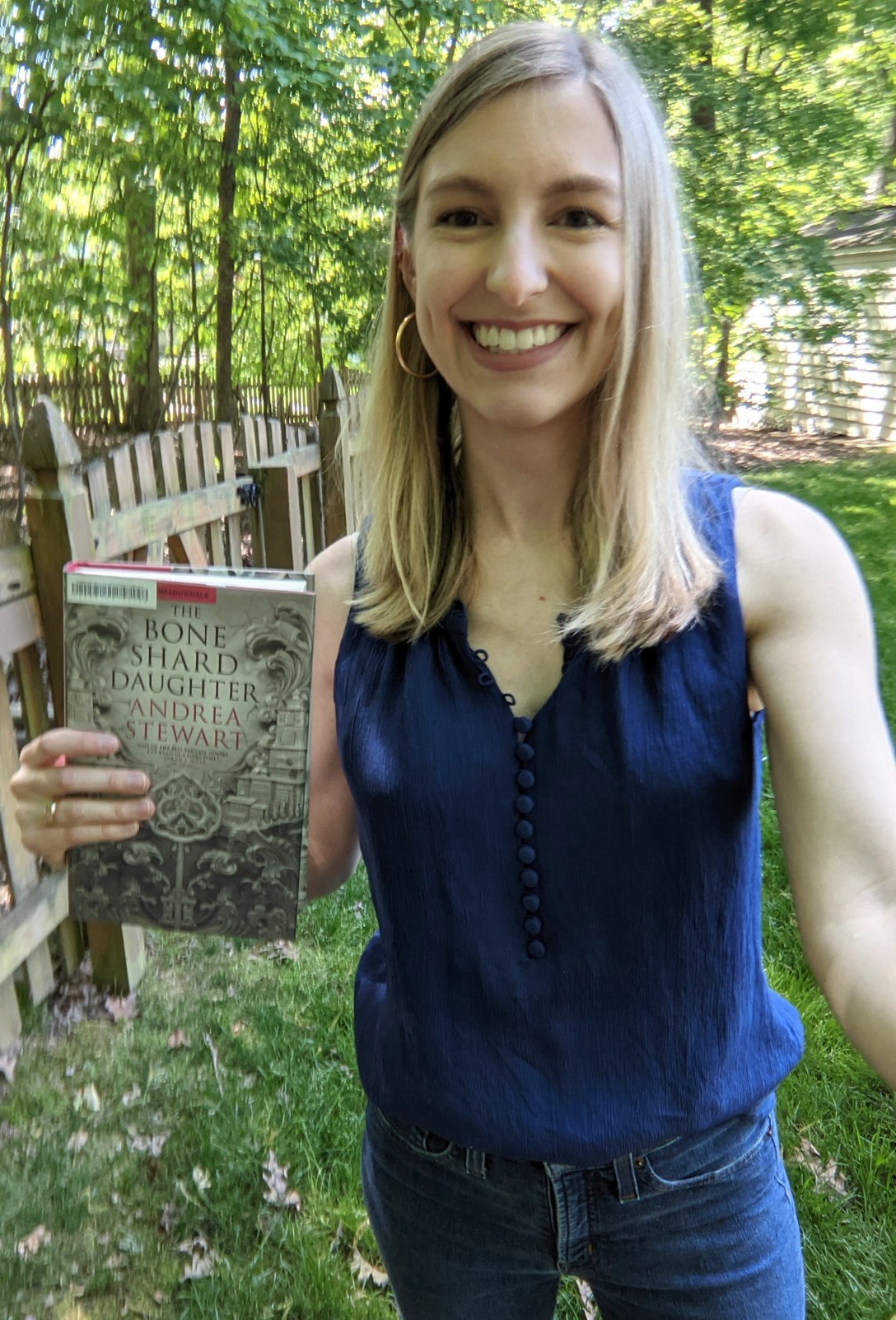 Alexis holds a library copy of The Bone Shard Daughter in front of a fence and trees.