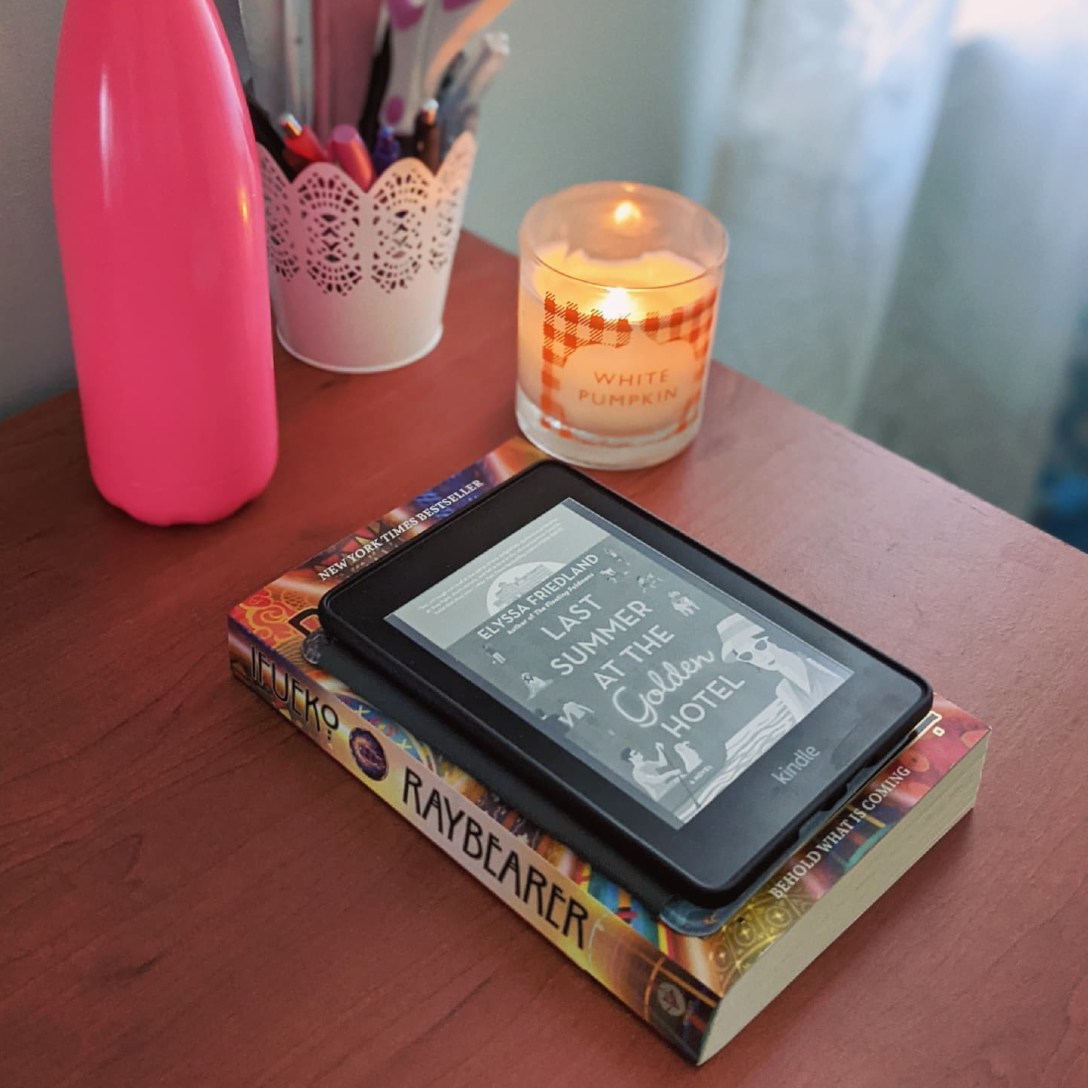 A kindle sits on top of a paperback copy of Raybearer on a desk. A lit white pumpkin candle sits behind it, next to a cup of pens and office supplies and a pink water bottle.