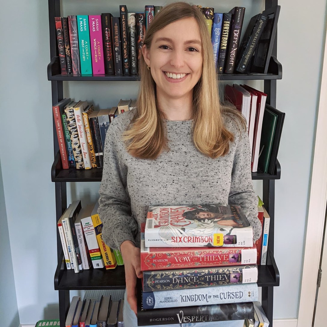 Alexis. wearing a gray sweater, stands in front of a bookshelf while holding a stack of books.