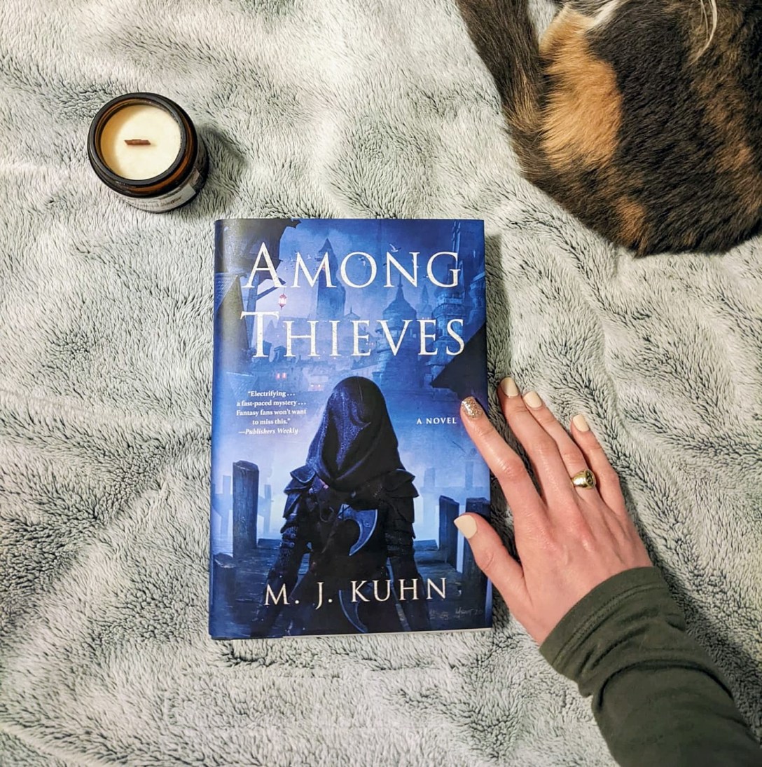 A copy of Among Thieves rests on a gray blanket next to a candle and a dilute calico cat.