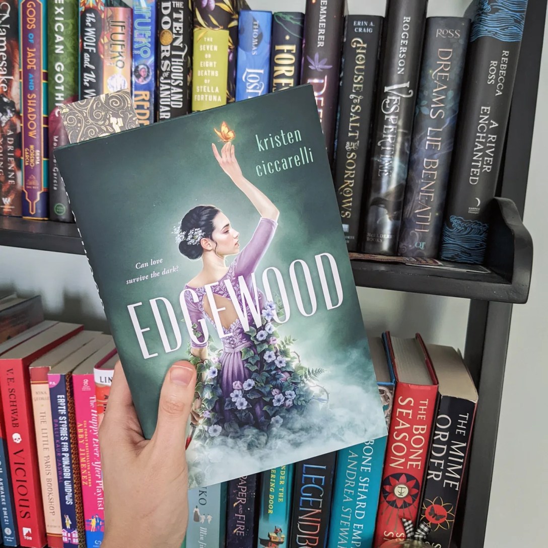 Alexis holds a copy of Edgewood- green with a girl wearing a purple dress and holding a dagger- in front of a bookshelf.