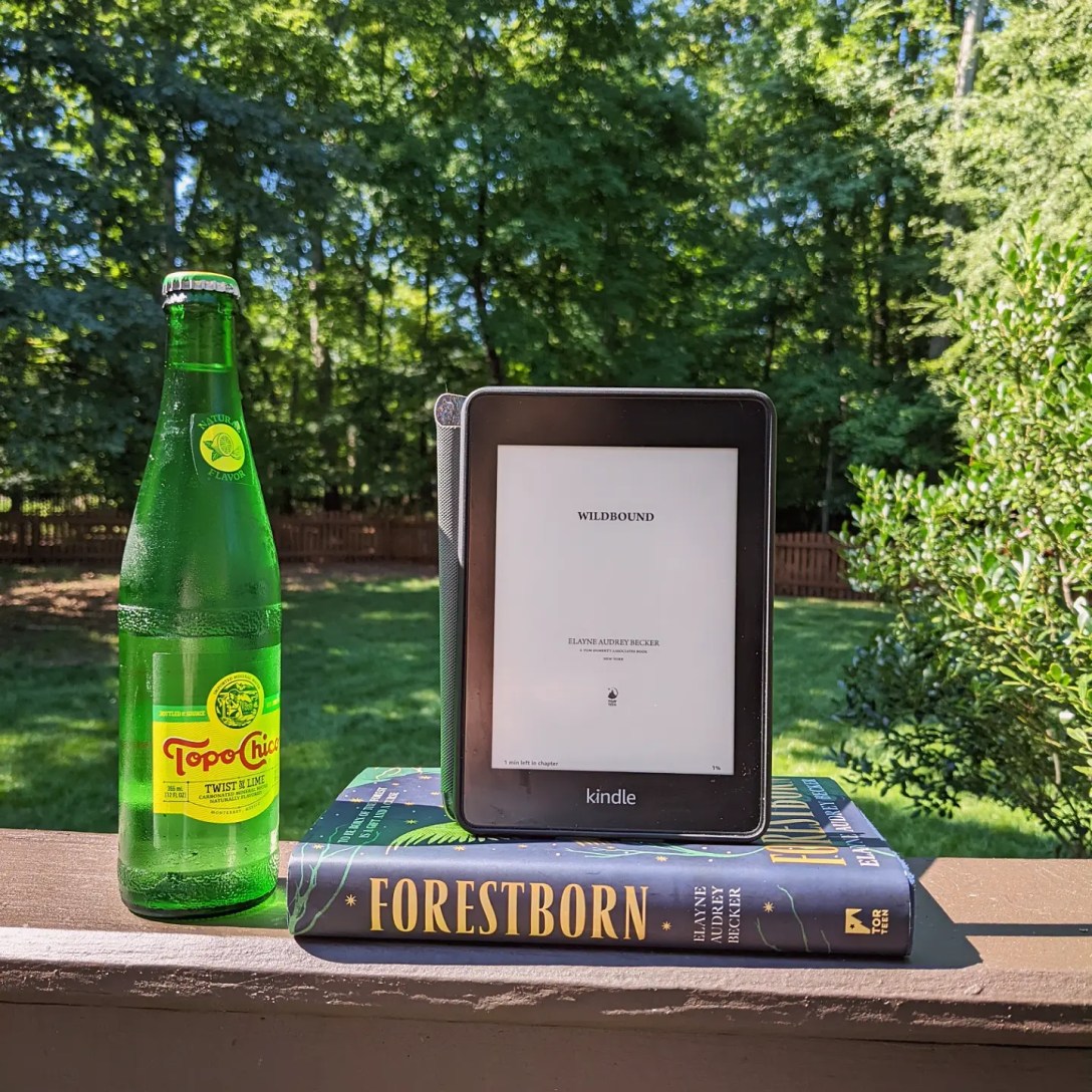 An e-ARC of Wildbound on a Kindle sits on top of the spine of Foresborn in front of a green forest. A bottle of Topo Chico sits on its left.