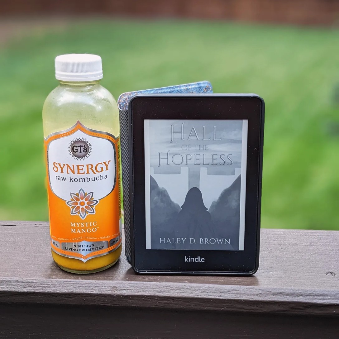 On a wooden railing, a bottle of mango kombucha rests next to a Kindle in front of green grass