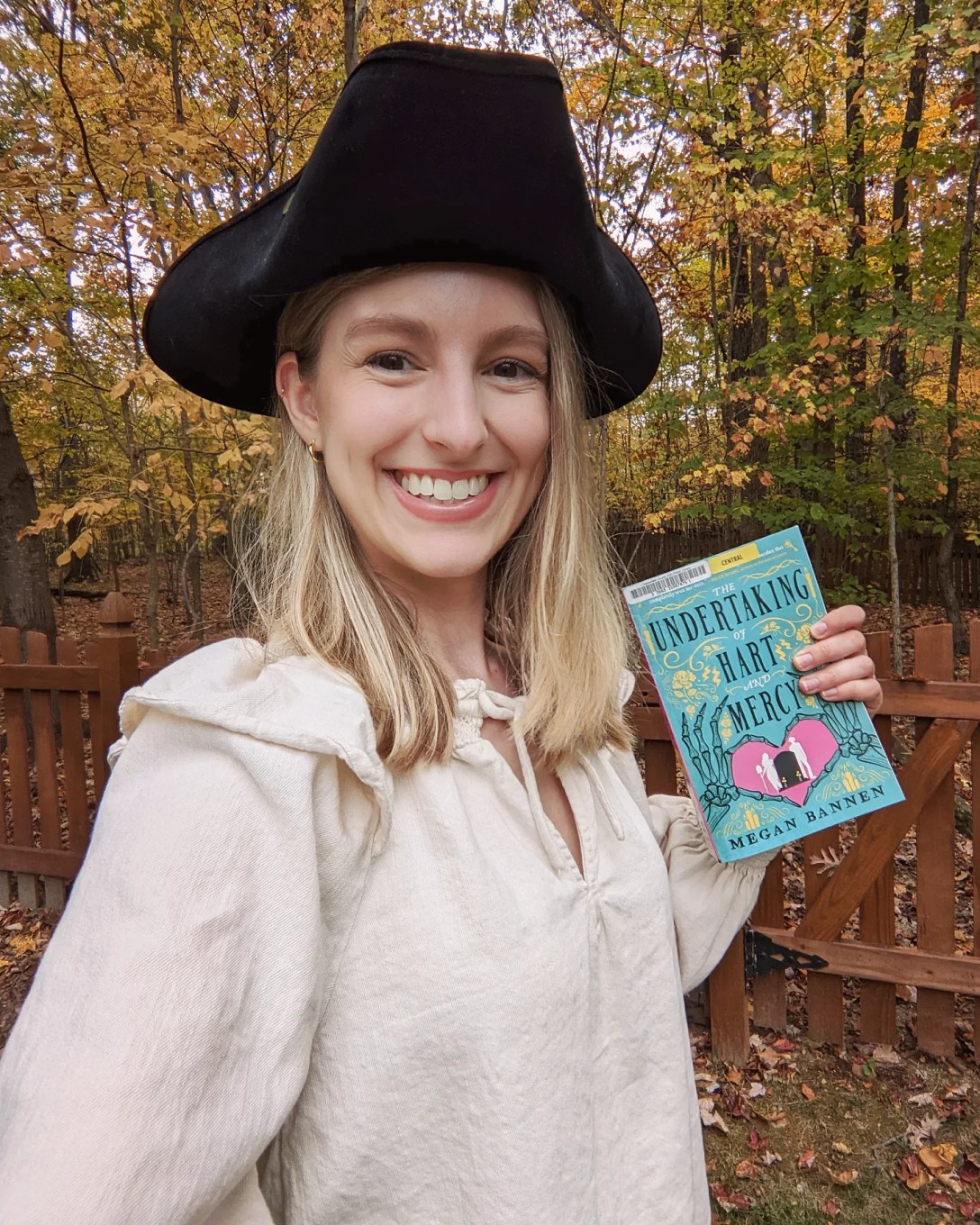 Alexis wears a pirate Halloween costume and holds a copy of The Undertaking of Hart and Mercy in front of fall foliage.