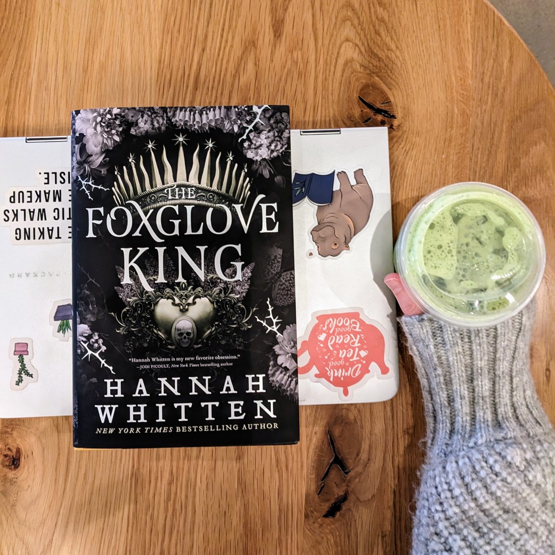 The Foxglove King sits on top of a laptop on a table at a coffee shop, next to Alexis's hand in a gray sweater, which holds an iced matcha latte.