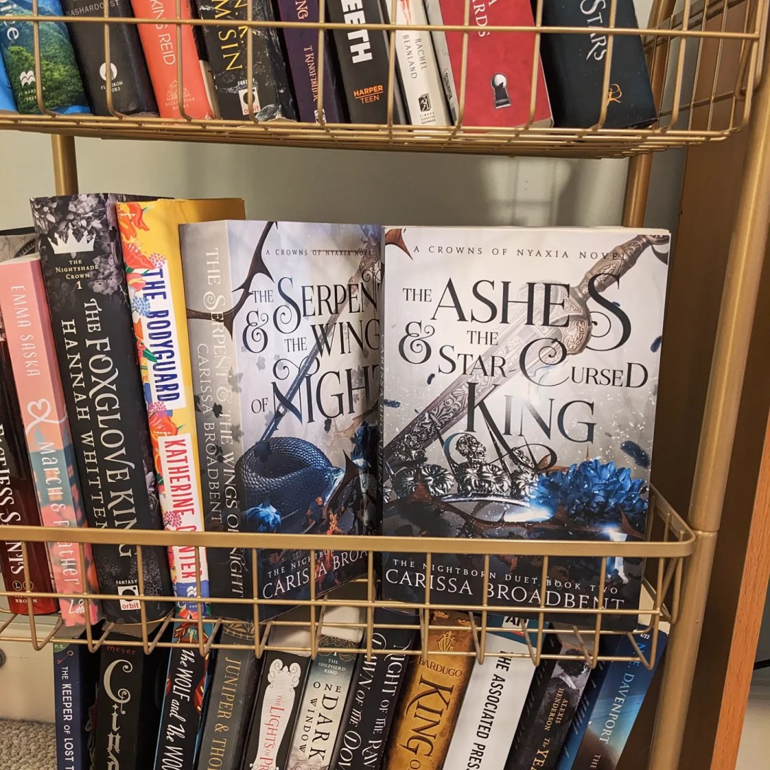 A paperback copy sits in a gold book cart next to the first book.