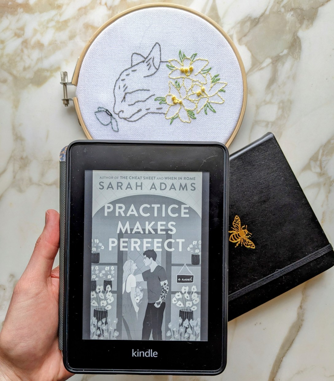 Alexis' hand holds her Kindle over a marble table with a black notebook with a honeybee and an embroidery with a cat and yellow flowers