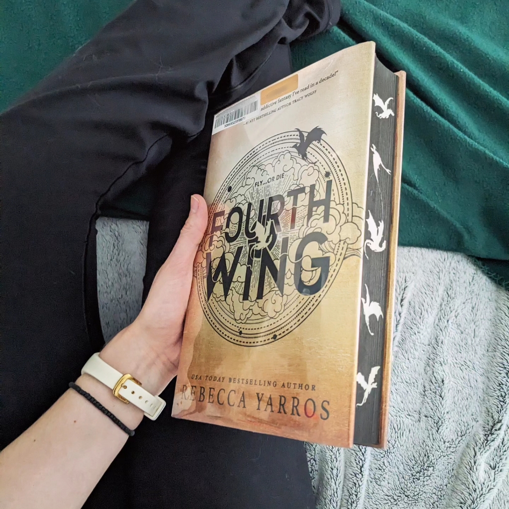 Alexis' hand holds a library copy of Fourth Wing over a gray blanket and a green blanket
