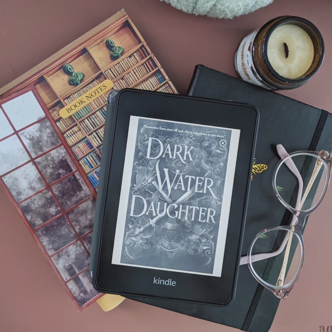A Kindle ARC copy of Dark Water Daughter lays on a desk on 2 notebooks and next to a pair of pinkish-clear glasses and a candle