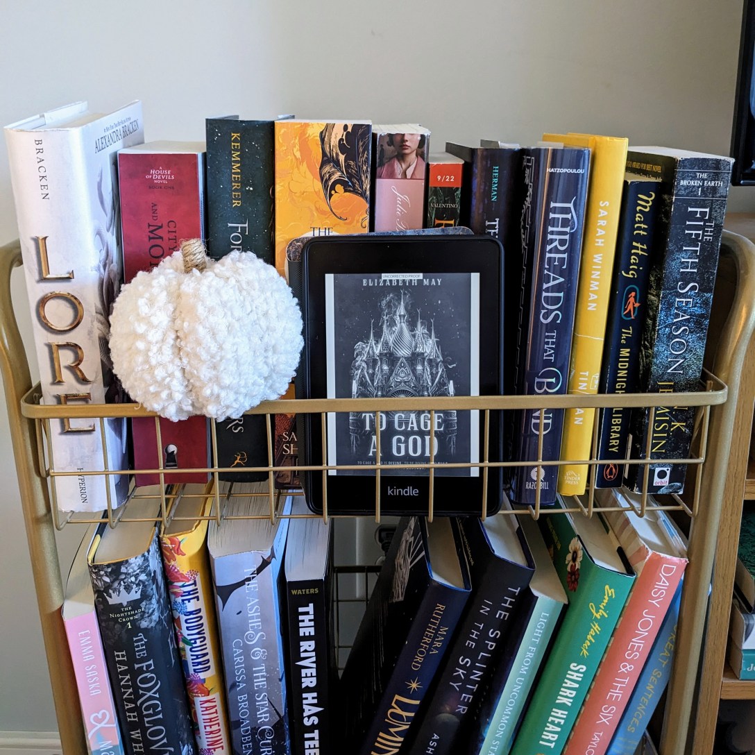 A Kindle with To Cage A God sits on a gold book cart next to a stuffed pumpkin