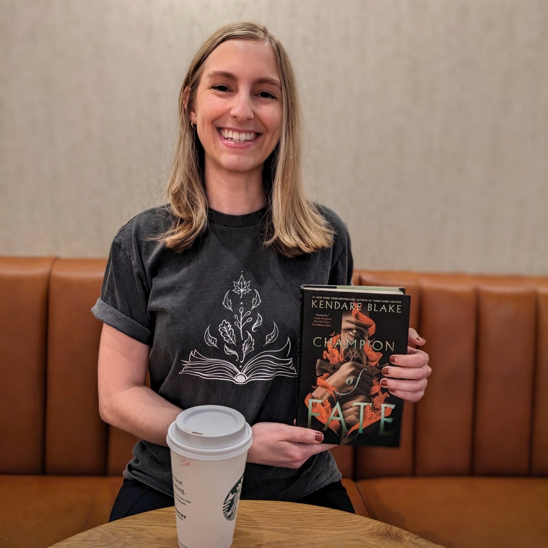 Alexis sits in a coffee shop and holds Champion of Fate in her hands.