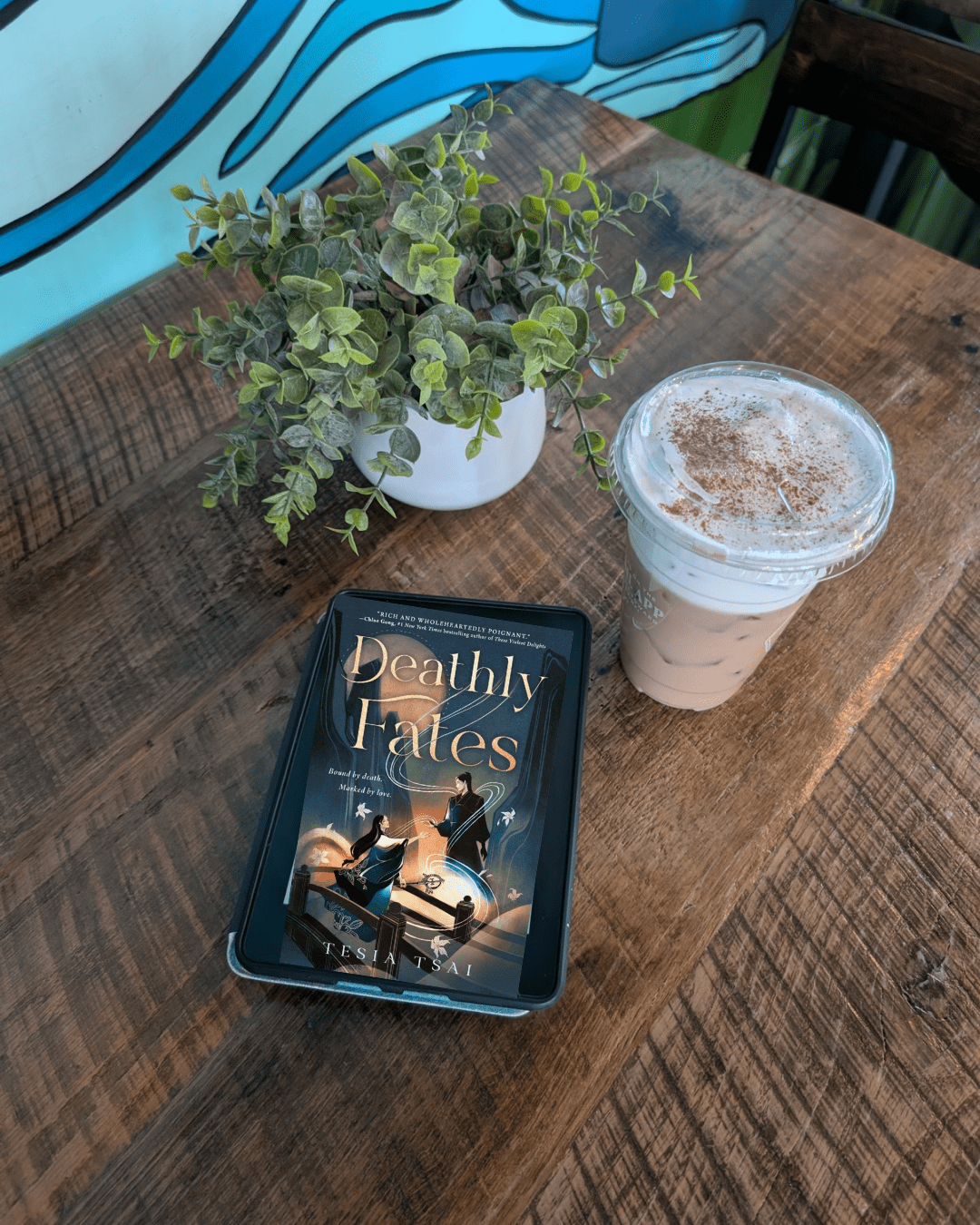 Kindle with Deathly Fates cover, a plant, and a chai latte on a coffee shop table